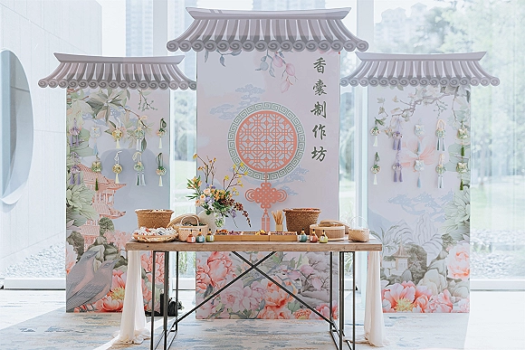 Wedding favor table with hanging tassel favors and wedding favors display on a wooden draped table beside floor-to-ceiling windows and greenery outside
