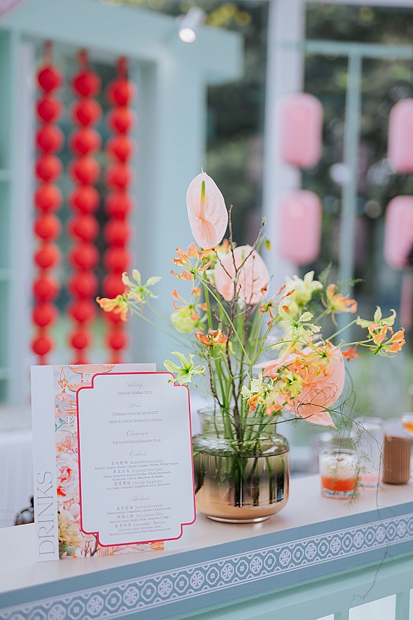 Wedding drink station with wedding bar menu sign, anthurium centerpiece in a glass vase, and cocktail glasses under hanging lanterns outdoors