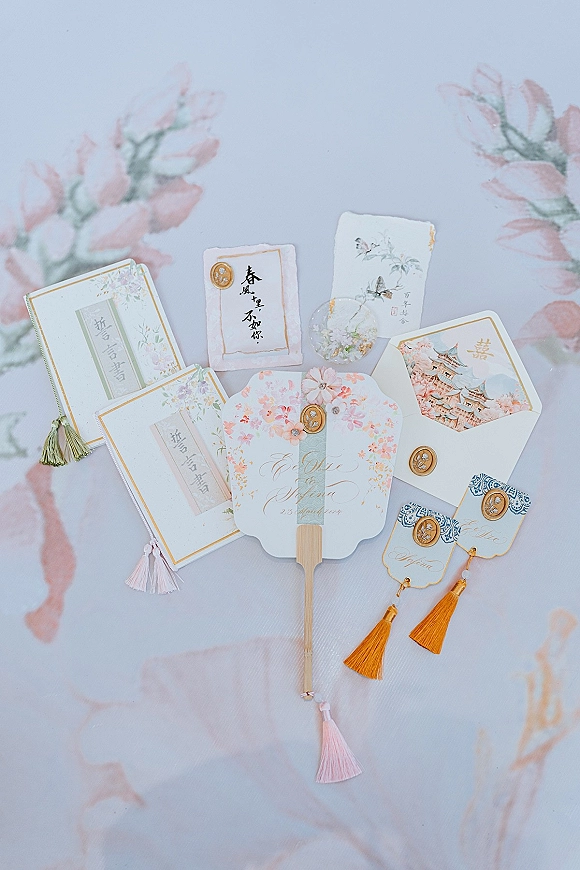Invitation suite flatlay with wax seals, tassels, and gold foil lettering on pastel surface with painted floral backdrop