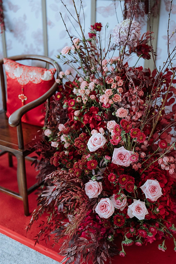 Wedding floral arrangement of pink roses and hydrangea with flowering branches beside a red velvet chair against floral wallpaper