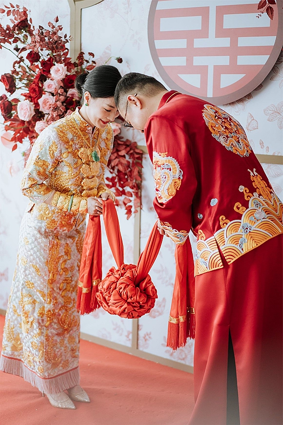 Tea ceremony moment as Chinese tea ceremony couple bow in red and gold attire with jade jewelry before a double happiness backdrop