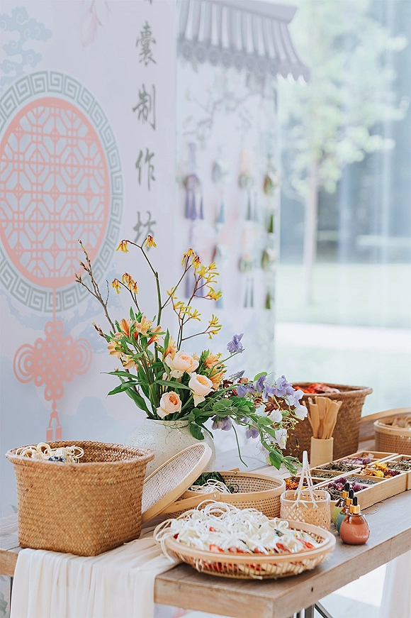 Wedding favor table with woven baskets and rattan trays, pastel flowers in a ceramic vase, and a backdrop sign by window light