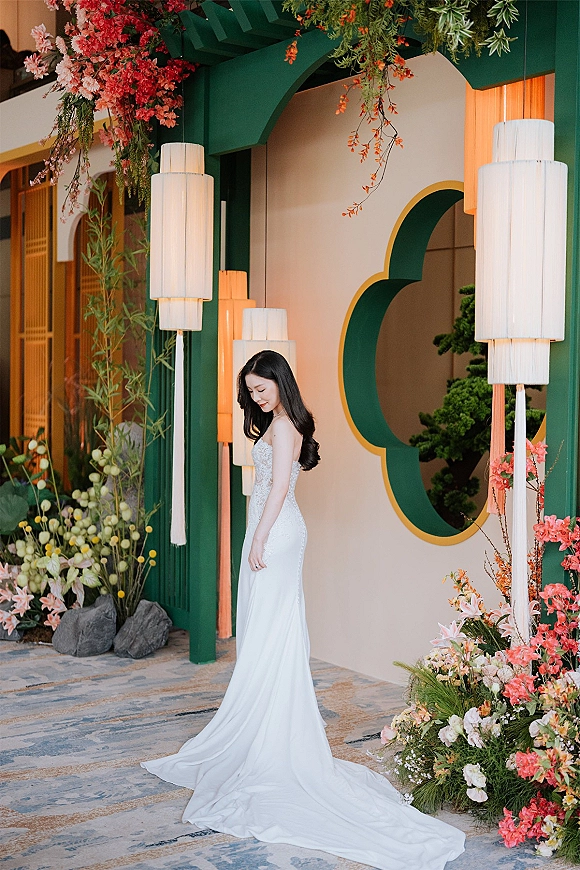 Bridal portrait of a bride in a strapless lace wedding dress with a long train, looking down by a floral arch with lanterns indoors