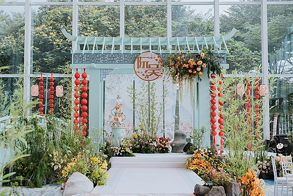 Ceremony backdrop with a wedding ceremony arch in pagoda style, hanging red lanterns and floral greenery beside a white aisle runner in a glasshouse garden setting