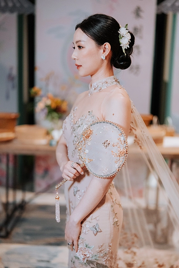 Bridal portrait of a bride holding hand fan in a high neck embellished dress and veil, side profile in soft window light indoors