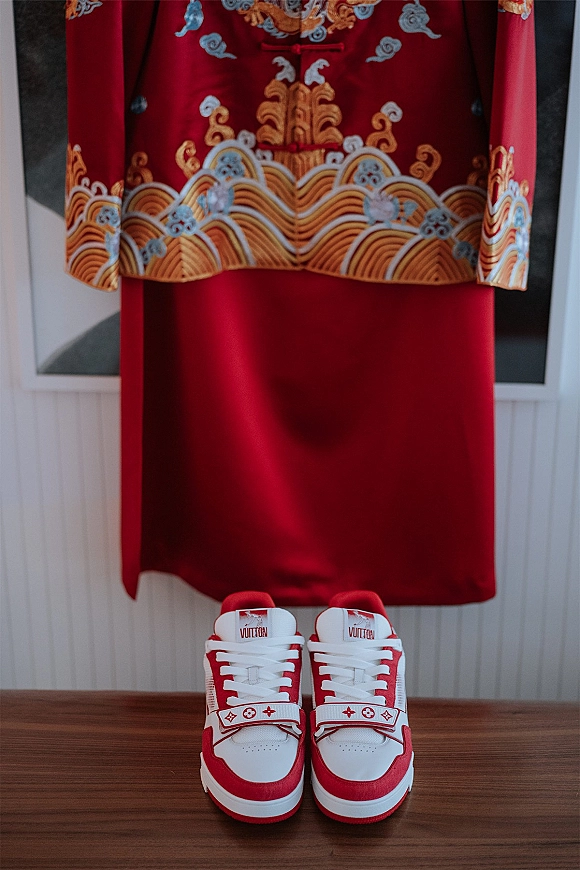 Groom wedding outfit flat lay with red embroidered jacket, matching trousers, and red-and-white sneakers on a wood tabletop by windows