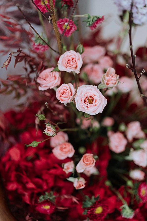 Wedding florals blush rose arrangement with red blooms, buds, branches and lush greenery against a soft indoor backdrop