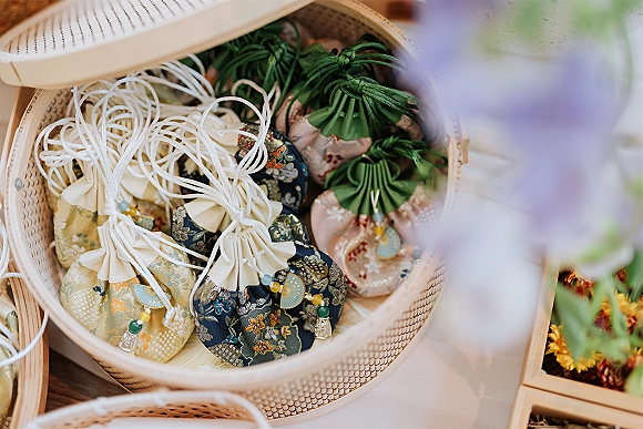 Wedding favors in fabric pouch wedding favors with drawstring tassels and beaded charms arranged in a woven basket on a tabletop with blurred flowers