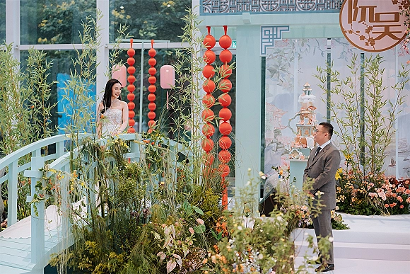 Ceremony moment as bride enters in strapless gown toward groom in gray suit, framed by bamboo and red lanterns in a glasshouse venue