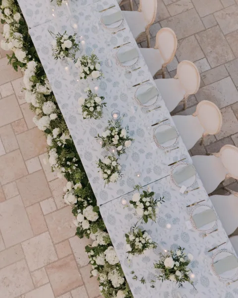 Reception tablescape with long table wedding decor featuring white rose garland, bud vases, candles, gold flatware on a stone patio