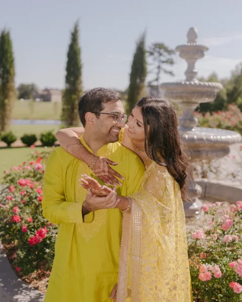 Couple portrait with mehndi couple photo, in yellow traditional outfits hugging by a stone fountain amid pink flowers and greenery
