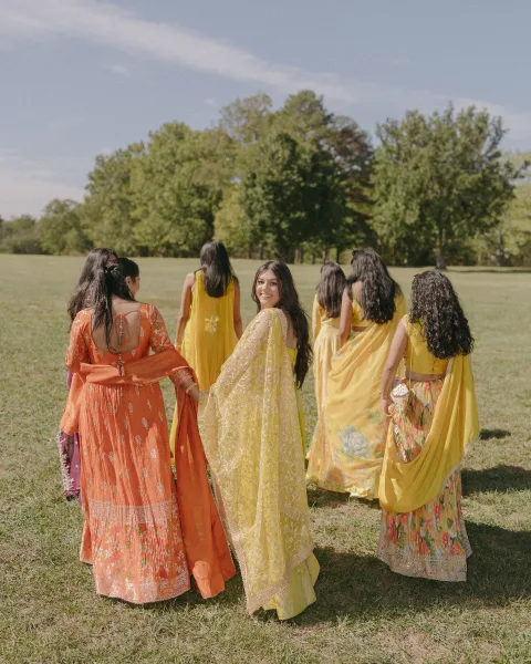 Bridesmaid group photo of South Asian bridesmaids in yellow sarees and an orange lehenga walking in sunlight across a grassy field