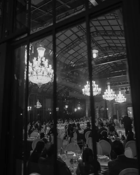 Wedding reception decor with crystal chandeliers and candle centerpieces over round banquet tables in a brick-walled ballroom with arched glass ceiling
