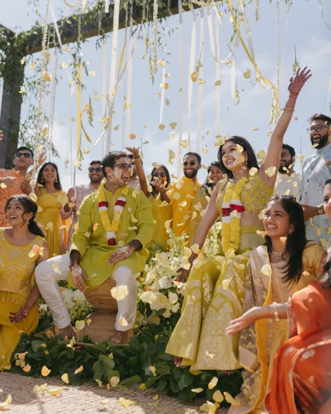 Haldi ceremony couple in a yellow lehenga and green kurta under floral garlands, with petal confetti and ribbons in an outdoor setting