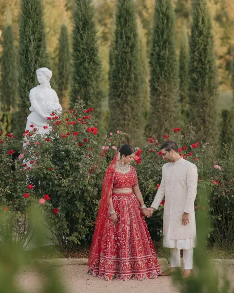 Couple portrait of an Indian wedding couple holding hands, bride in a red lehenga, walking along a rose garden path with statues