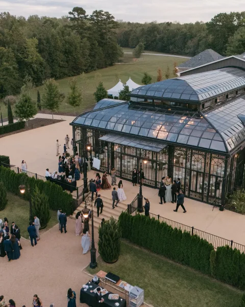 Wedding reception venue with glass conservatory and black iron framework, patio cocktail tables and bar setup beside a white tent on lawn
