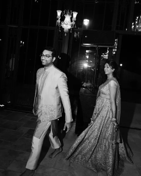 Wedding entrance of bride and groom in embroidered lehenga and sherwani, dupatta flowing, walking into a dark venue under a chandelier