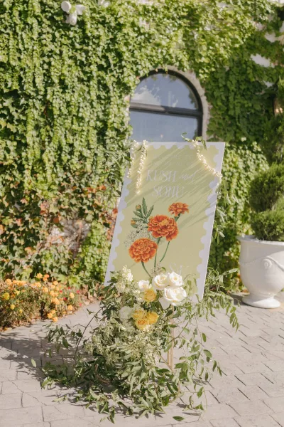 Wedding welcome sign on an easel with an acrylic floral illustration and a greenery garland, set against an ivy-covered wall with arched window