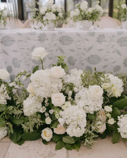 Wedding floral arrangement with white rose centerpiece and hydrangeas, greenery garland and planter boxes on a patterned tablecloth by glass windows