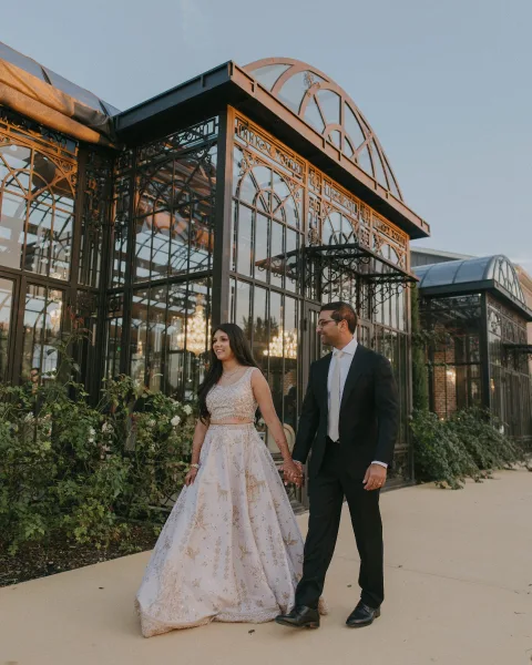 Couple portrait of bride and groom holding hands, bride in sequin lehenga with henna, walking by a glass conservatory at dusk