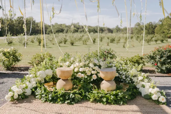 Wedding ceremony backdrop with hanging ribbons and white rose florals over a woven rug, set along a gravel path in an orchard field