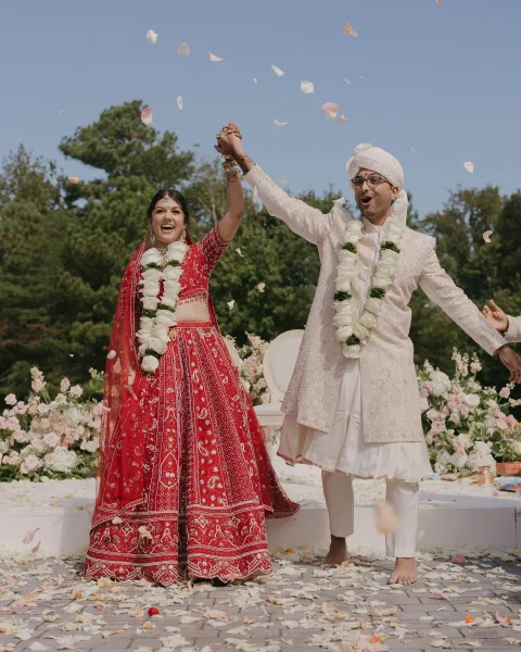 Wedding recessional of newlyweds celebrating with raised hands, rose petals falling as they stand on a floral stage in a garden