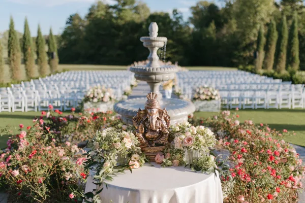 Outdoor ceremony setup with garden wedding ceremony rows of white folding chairs facing a stone fountain, rose and greenery aisle florals under blue sky