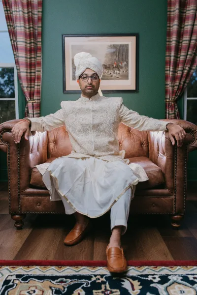Groom portrait in embroidered sherwani and turban, wearing glasses while seated on a leather sofa against a green wall with windows