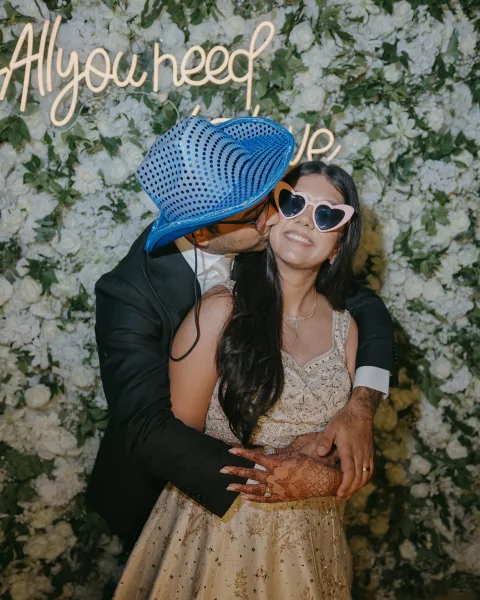 Wedding couple portrait with groom kissing bride’s cheek, wearing heart sunglasses before a white flower wall and neon sign backdrop