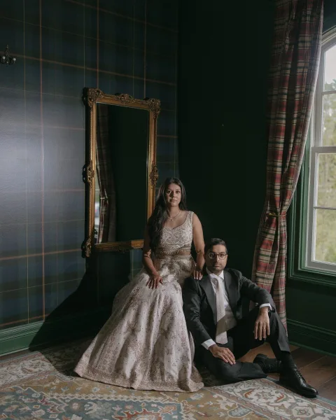 Couple portrait of a South Asian bride in a bridal lehenga beside her groom in a gray suit with glasses, posed by an ornate gold mirror in window light