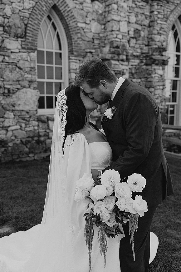 Wedding kiss portrait of bride and groom kissing, her veil flowing as she holds a bouquet on a lawn before a stone building with arched windows
