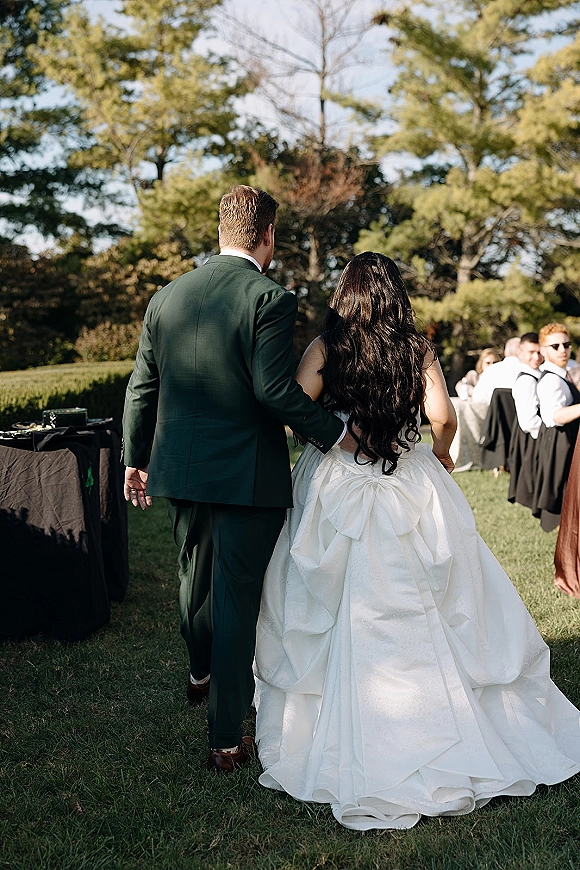 Wedding couple portrait of bride in a white ballgown and groom in a tuxedo walking away past black cocktail tables on a sunny lawn