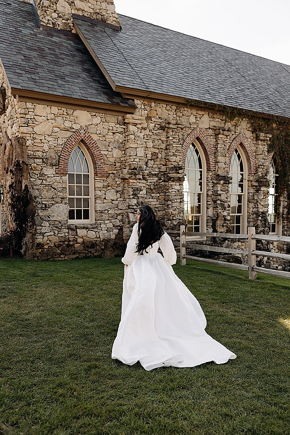 Bridal portrait of a bride from behind in a long sleeve wedding dress with a train, walking on grass beside a stone church with arched windows