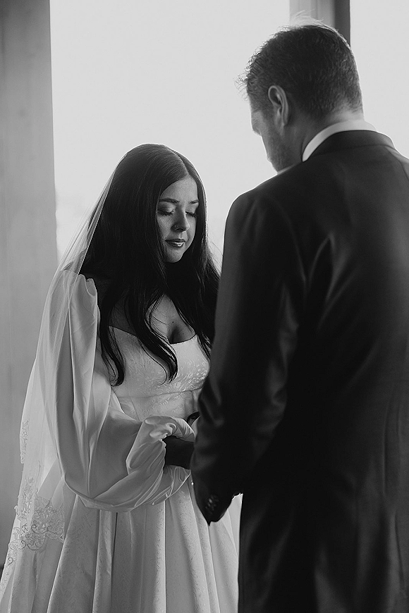 Ceremony moment as bride and groom hold hands during wedding vows, bride in veil and long-sleeve dress in soft window light indoors