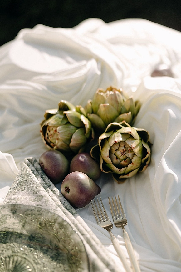 Reception tablescape with wedding place setting on white linen, featuring artichokes and plums beside forks and a glass charger plate against dark backdrop