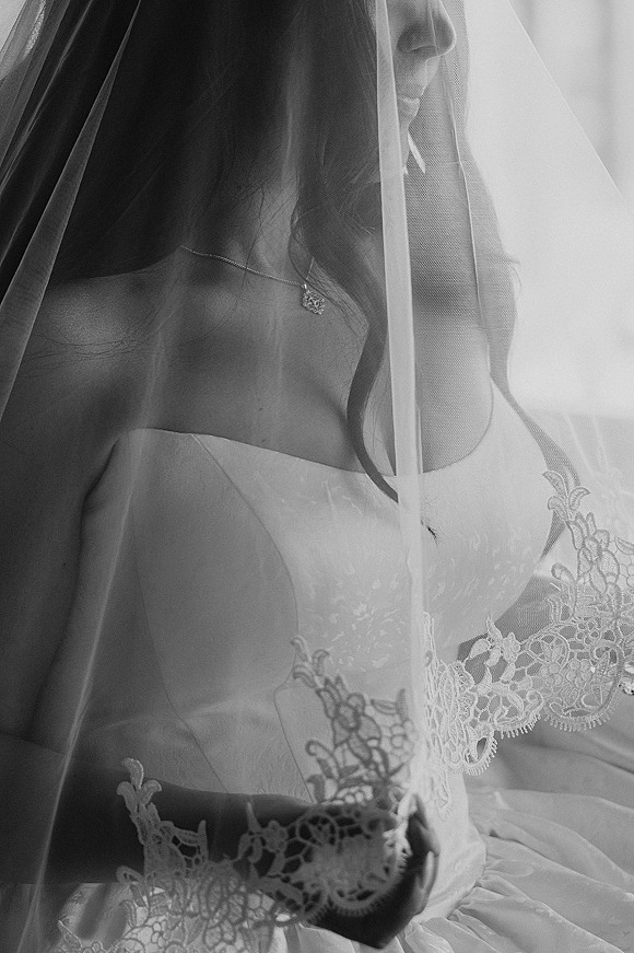 Bridal portrait of a bride under veil, looking down in a strapless lace-trimmed dress with a pendant necklace in bright window light