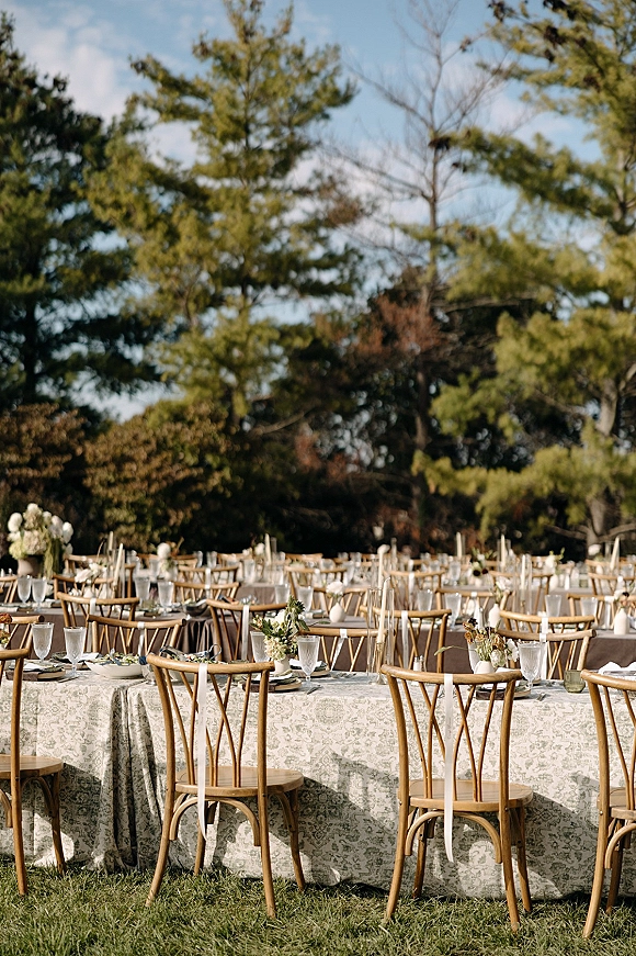 Reception tablescape for an outdoor wedding reception with patterned tablecloth, wood crossback chairs, taper candles and floral vases on a lawn under trees