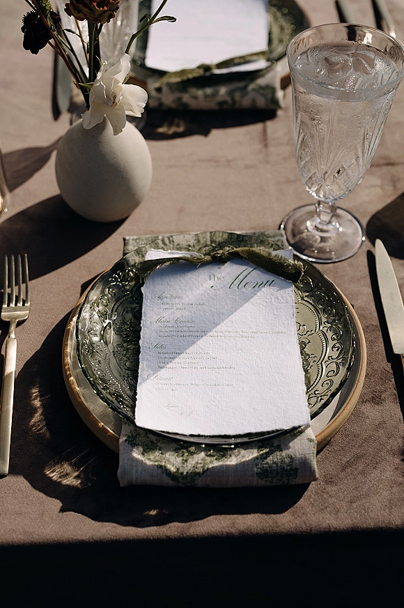 Reception tablescape with wedding place setting featuring a menu card on layered plates, green glass charger, gold flatware, and bud vase on taupe linen in sunlit shadows