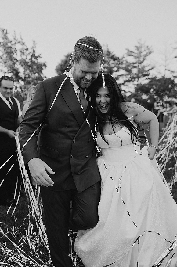 Recessional moment as bride and groom laugh walking down the aisle with metallic streamers, guests cheering on a tree-lined lawn