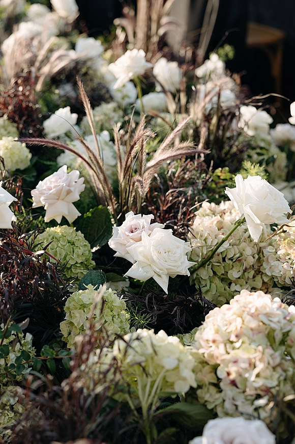 Wedding floral arrangement of white roses and hydrangea blooms with greenery accent in a sunlit garden aisle with blurred foliage