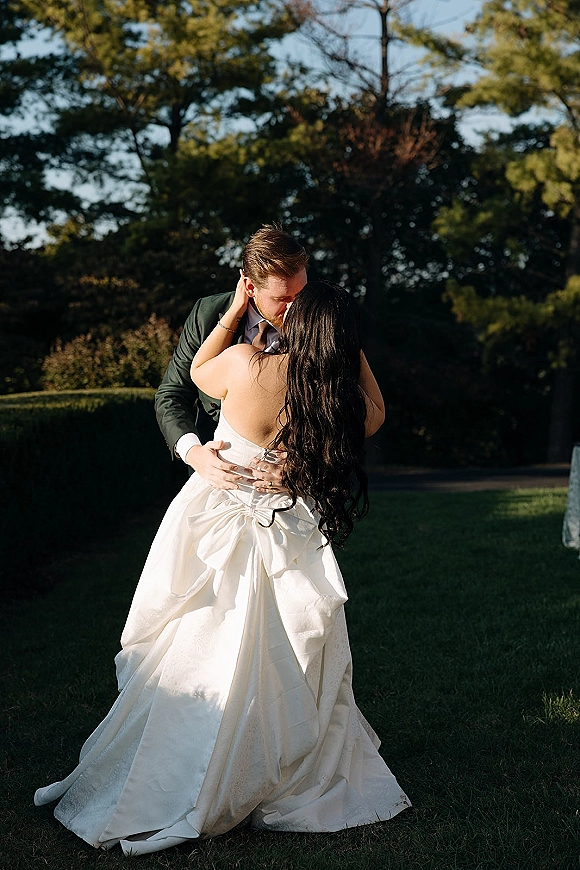 Wedding kiss portrait of bride in a strapless wedding dress and groom in suit dipping into a kiss on a sunny lawn with trees and hedges