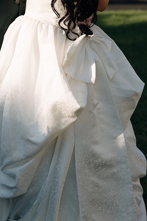 Wedding dress close-up showing an oversized bow on textured satin jacquard gown, with bride’s curls and groom’s black suit on a lawn