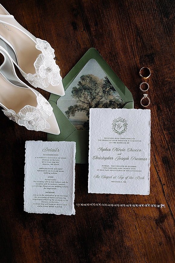 Invitation suite with deckle edge wedding invitation and green envelope, styled with rings and lace bridal shoes on a dark wood table