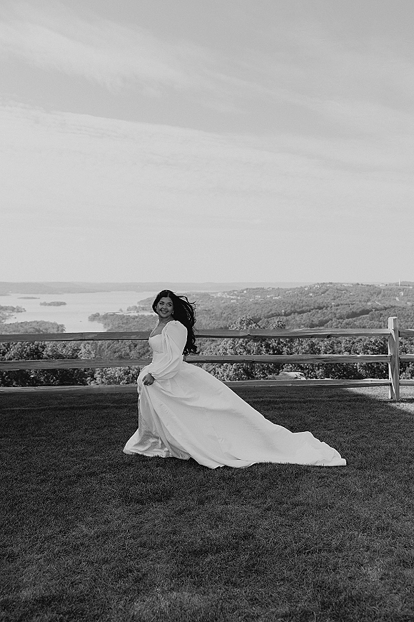 Bridal portrait of a bride twirling in a long sleeve wedding dress with a flowing train on grass by a lake and rolling hills