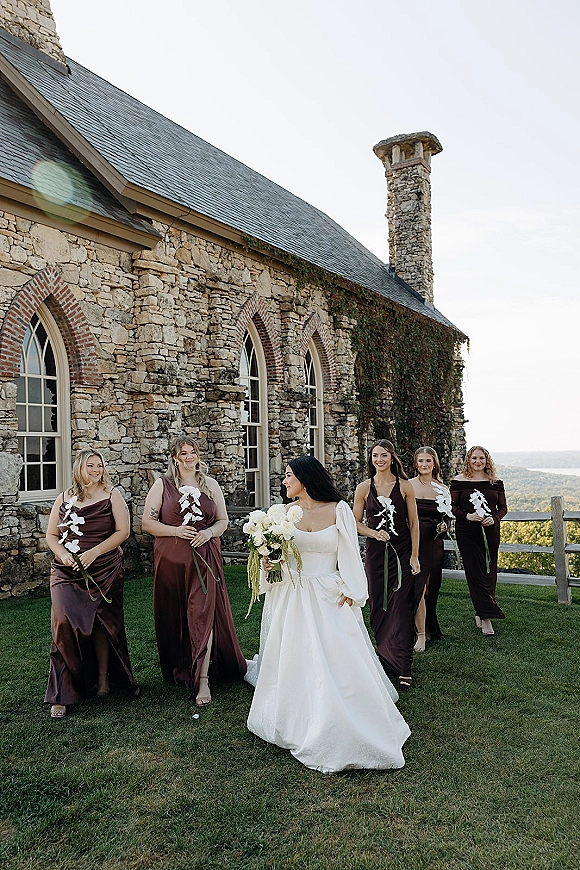 Bridesmaid group photo with bride and bridesmaids in burgundy and brown dresses holding calla lily bouquets by an ivy stone building