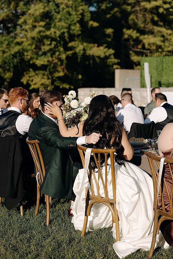 Reception moment at outdoor wedding reception as bride in a white gown touches groom’s face while seated at a garden table with guests behind