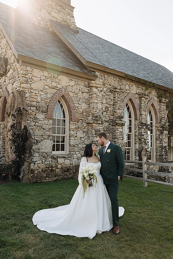 Couple portrait of bride holding a white flower bouquet and groom in suit by a stone church with arched windows and sun flare
