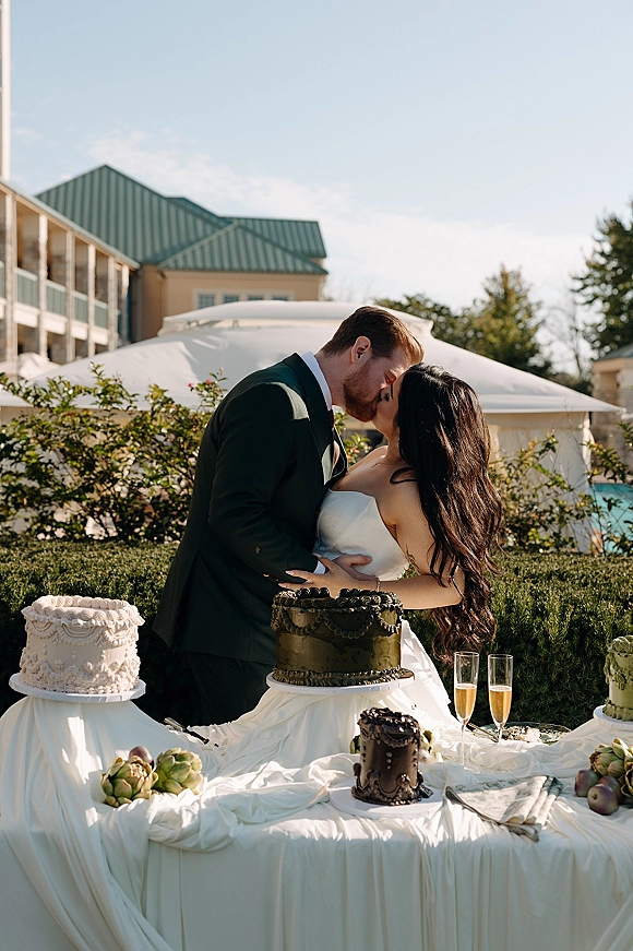 Wedding kiss portrait of bride and groom kissing beside a cake cutting table with champagne flutes under a white canopy tent outdoors