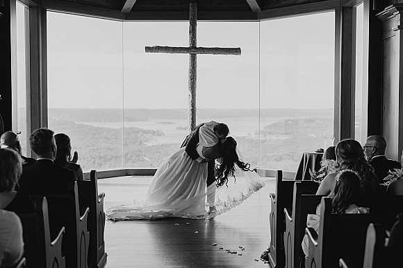 Wedding kiss at the altar as the groom dips the bride in her veil and dress, wooden cross behind, guests in a chapel with mountain-view windows