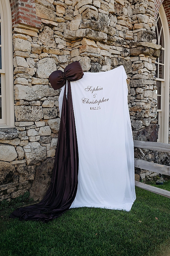 Wedding welcome sign with draped fabric and oversized bow, featuring calligraphy lettering against a stone wall by a window on lawn